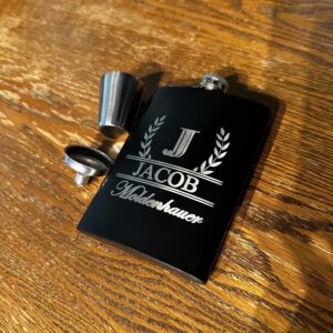 A black flask beside a shot glass and funnel on a wood background. The flask is engraved with a letter and a name.