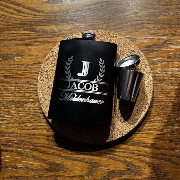 A black flask beside a shot glass and funnel on a wood background. The flask is engraved with a letter and a name.